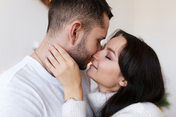 The concept of new year's love story. A guy and a girl in white cozy sweaters hugging, kissing, dancing and fooling around on the background of a white fireplace, decorative Christmas tree and bokeh.