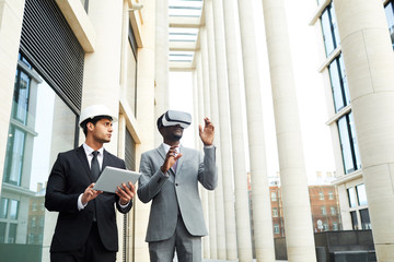 Young businessman in hardhat and suit using tablet pc and looking at his colleague while he wearing virtual reality goggles and walking in the city