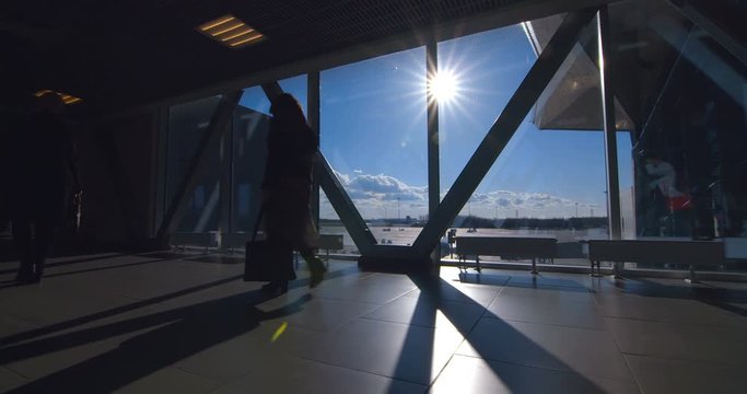 Passengers go through the glass passage where the runway with planes and other infrastructure is visible from the windows. The bright sun shines through the window and only the silhouettes of passenge