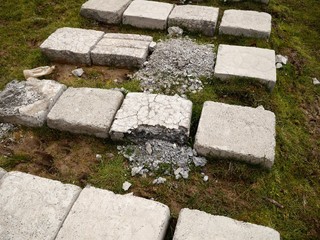 Photo of a path paved with rows of grey concrete blocks, with some of the blocks ruined. Nature vs artificial constructions concept. Road for walking on a lawn.