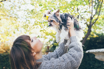 Lifestyle photo in the autumn park of a beautiful happy woman who is holding her cute little biewer terrier dog with stylish fur-cut and nice bow. Owner with the dog. Natural light, selective focus.