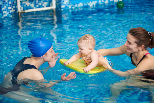 Adorable baby enjoying swimming on floating board in pool with his mother and trainer helping. Side view. Interesting game and useful pastime for children. Newborn baby at sportive active family