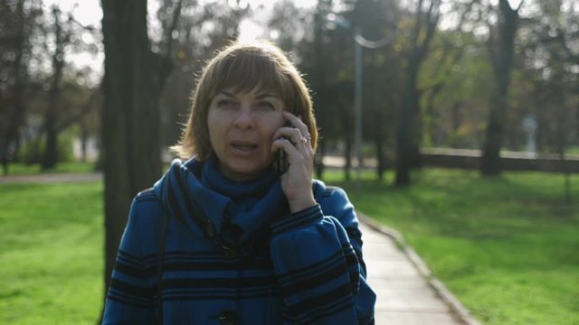 Cheery blonde woman strolling and talking on phone in a park in summer in slo-mo 