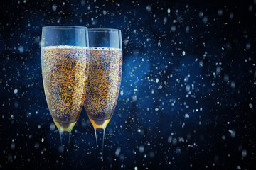 Two elegant romantic glasses with sparkling golden champagne against a background of blue blurry lights with snow swirling around them. Christmas, New Year, holiday background. Greeting card template.