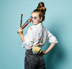 Young artist woman with a set of brushes in shirt with paint stains is at work, standing back, looking over her shoulder
