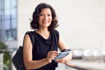 Portrait Of Mature Businesswoman With Passport In Airport Departure Lounge Using Mobile Phone