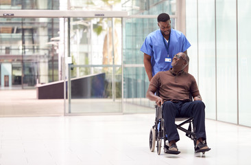 Male Nurse Wearing Scrubs Wheeling Patient In Wheelchair Through Lobby Of Modern Hospital Building - Powered by Adobe