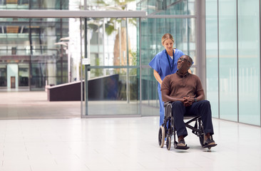 Female Nurse Wearing Scrubs Wheeling Patient In Wheelchair Through Lobby Of Modern Hospital Building