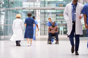 Fotobehang Female Nurse Wearing Scrubs Wheeling Patient In Wheelchair Through Lobby Of Modern Hospital Building © Monkey Business