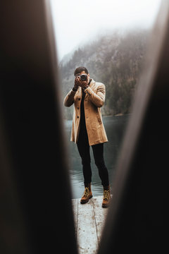 Full body shot of young male photographer in brown coat taking pictures with a small mirrorless camera standing on a lake house deck in lake plansee, Austria
