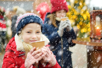 Two adorable sisters drinking hot chocolate on traditional Christmas fair in Riga, Latvia. Children enjoying sweets, candies and gingerbread on Xmas market.