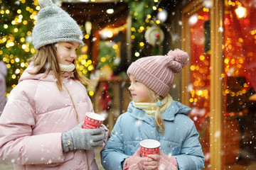 Two adorable sisters drinking hot chocolate on traditional Christmas fair in Riga, Latvia. Children enjoying sweets, candies and gingerbread on Xmas market.