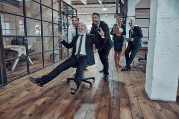 Obraz premium Playful businessmen pushing their boss on the office chair while running in the hallway