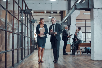 Full length of two colleagues in elegant formalwear discussing business while walking through office hall