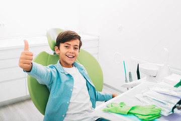 Happy little boy showing thumbs up . He sitting at dental chair, after teeth treatment. Yes, my teeth is healthy