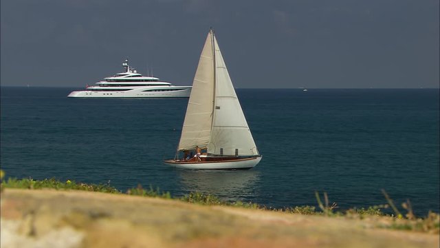 Medium low-angle panning shot of a small sailing boat with sightseeing tourists, and a stationary luxurious yacht at the sea, Vauban  Port Harbour, France