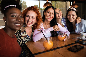 Group of young female friends having fun in cafe, talking and laughing while sitting at table and making selfie.