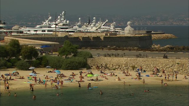 Medium panoramic high-angle still shot of tourists at a crowded beach, Nomade sculpture facing the sea, and Boats docked at the harbour, Port Vauban, Antibes, France