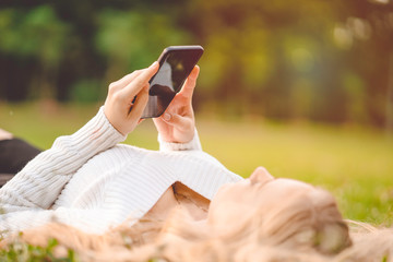 Young caucasian woman lying on a lawn in a park, holding smartphone in hands and using to search website on screen, chatting message and playing social media for relaxation, feel happiness and freedom