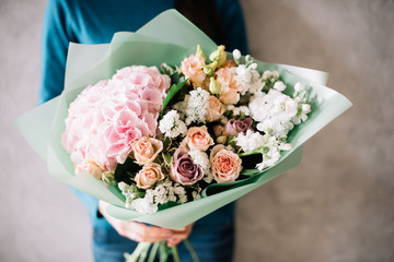 very nice young woman in a blue sweater holding a beautiful blossoming flower bouquet of fresh hydrangea, roses, carnations, matthiola, in pink and pastel cream colors on the grey wall background © anastasianess