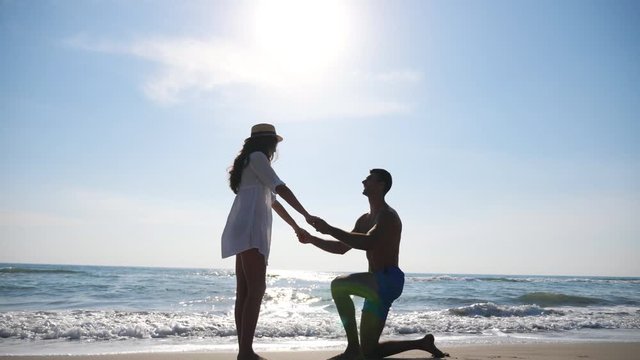 View on newlyweds at seascape background. Young man stand on knee and marriage proposal to his girlfriend summer vacation. Happy pair hugging and kissing on seashore. Concept of love and happiness