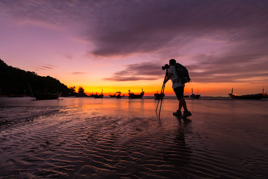 Silhouette of photographer taking a photo by using DSLR camera on tripod on the beach with sunset
