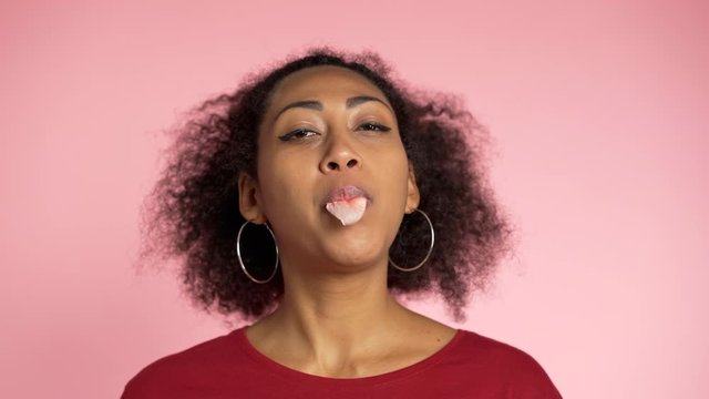 Playful girl in red wear blows bubblegum, chewing gum in slow motion. Pretty african american woman stands on pink background.