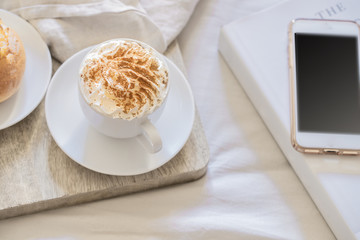 Cup of coffee, sweet bun, book and smartphone on the bed sheets. Good morning and relax at home concept.