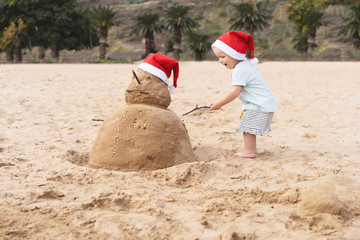 Little child in red santa hat with sand snowman on beach.