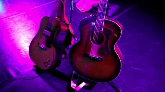 Acoustic guitars of dreadnaught and Southern Jumbo styles and a mandolin on guitar stands under violet colored light on stage before a show.