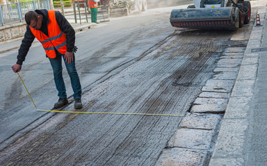 Worker measuring length on the road, polished with bitumen during repair work on the background of trucks with asphalt