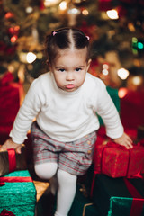 Stock photo portrait of adorable little Caucasian girl sitting in pile of red and green Christmas presents next to the New Year tree under snowfall.