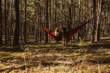 Two girls relaxing in the hammock hanging among the pine trees in the background. Autumn scenery, rays of light fall through the trees. One girl has short blond hair, the other has long brown hair.