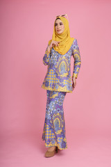 Beautiful female model wearing batik design "baju kurung" with yellow colored hijab, sitting on a chair isolated over pink background. Eidul fitri fashion and beauty concept.