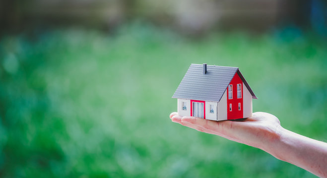 Adult Hand Is Holding Red House Model, Outdoors. Concept For New Home, Property And Estate