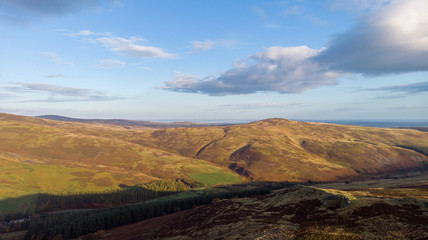 An aerial view of a mountain valley with grassy and stony slopes, trees and mountain range summits under a majestic blue sky and some white clouds