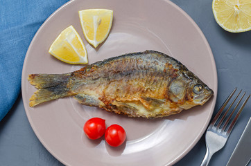 Fried appetizing fish crucian carp with seasoning on a beige plate on a gray background decorated with slices of tomato and lemon with a knife and fork. healthy eating concept.