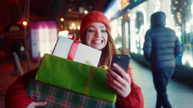 Ecstatic attractive woman in winter outfit carrying Christmas gift boxes using a smartphone walking in hurry on beautiful holiday street. Winter time. Happy New Year.