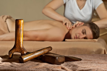 Blonde in a white T-shirt doing a massage to the guy. The man lies on the table on a yellow background. Wooden tools for Thai massage "Tok Sen" in the foreground. © Ivan