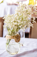 A bunch of baby's breath flowers in a glass jar on a table which is covered in a white tablecloth