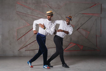 Two black funny guys in white shirts and straw hats are perky dancing against a textured wall.