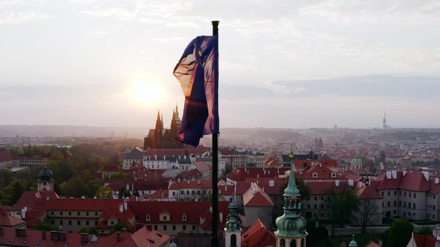 Aerial view town through waving flag on foreground. Lateral movement, drone shot, flying flag of EU in front of shining sun over scenic panorama Prague with red tiled roofs at sunrise