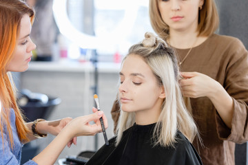 Women redhear make-up artist and hairdresser do hairstyle and stylish makeup for beautiful young blonde girl before start of fashion show. Colorful makeup design beauty and art salon concept