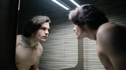 Young man in bathroom looking into a mirror at himself. He has a tattoo over his neck