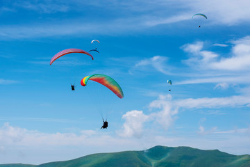 Paragliders, one by one, fly off the ground and ascend into the blue sky against the background of beautiful mountains. Excitement paragliding adventures in the sky