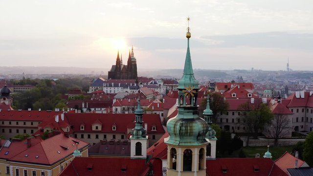 Aerial view over towers Lareta cathedral at sunrise. Lateral movement, shot from air, camera moving opposite belfries decorated symbol of christianity. Panorama above Prague and history buildings
