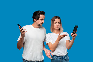 Brunet man in white tees and blue jeans is shocked looking at the smartphone screen of a blond woman