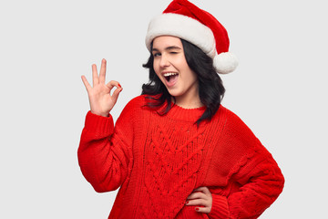 Excited brunette girl in a red sweater and Santa hat showing OK gesture and winking at the camera