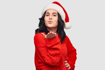 Excited brunette girl in a red sweater and Santa hat sending air kiss looking at the camera.