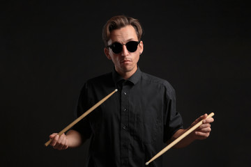 Stylish man drummer in a black shirt and sun glasses playing drums with sticks over dark background.